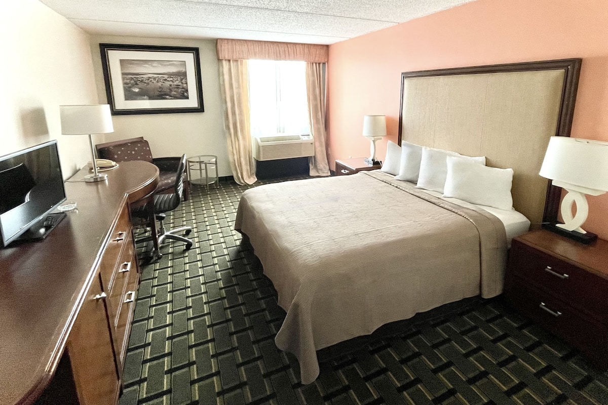 A hotel room with a bed, a desk with a computer, a chair, a TV, and a window with curtains.