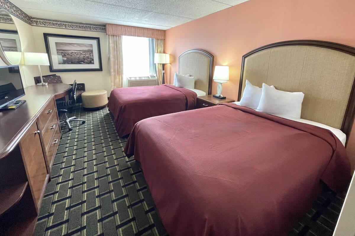 A hotel room with two beds, a desk with a computer, a chair, a lamp, and a picture on the wall.