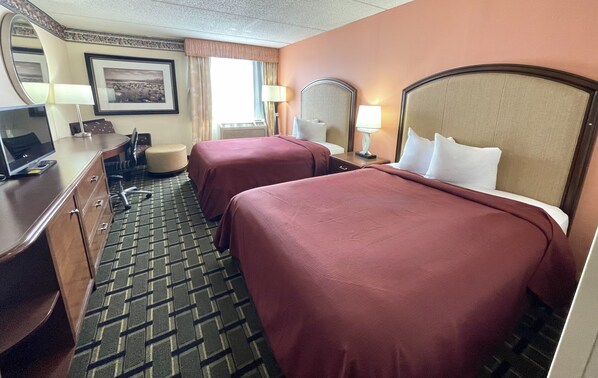 A hotel room with two beds, a desk with a computer, a chair, a lamp, and a picture on the wall.