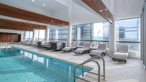 An indoor swimming pool with multiple lounge chairs and a view of a cityscape through large windows. - Sandman Signature Edmonton Downtown Hotel (Edmonton)