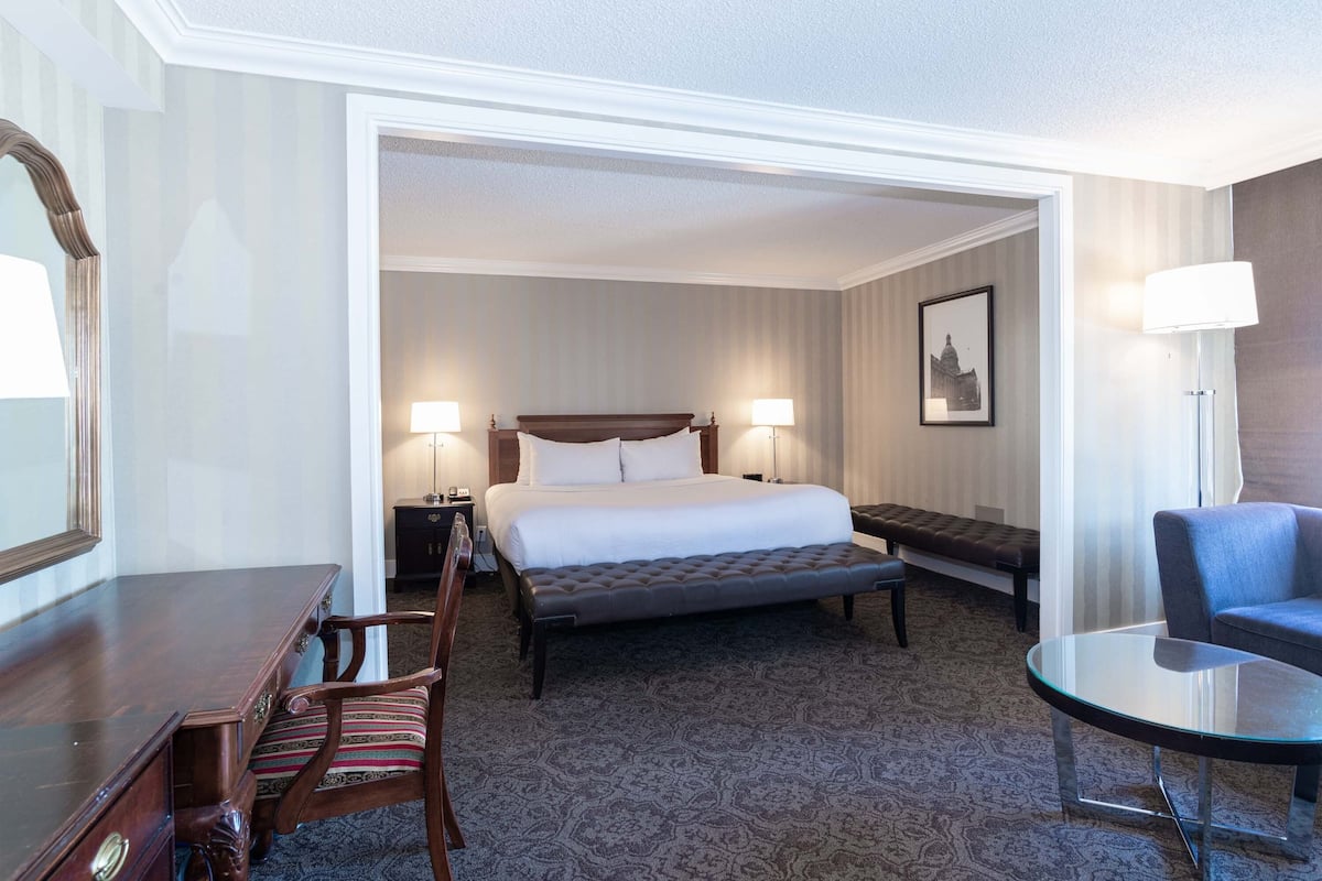 A hotel room with a large bed, a wooden desk, a chair, a small round table, and a bench.