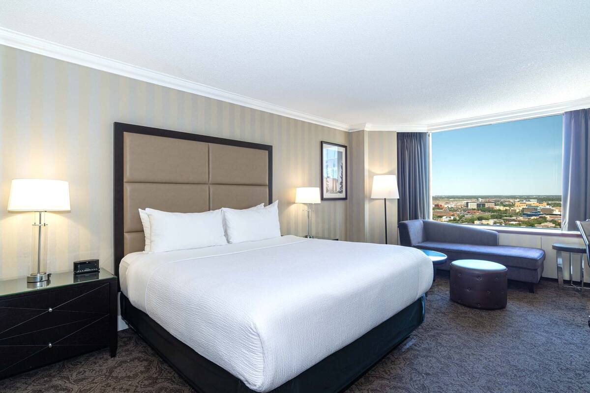 A hotel room with a large bed, two bedside tables with lamps, a sofa, and a view of the city.