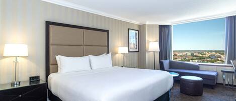 A hotel room with a large bed, two bedside tables with lamps, a sofa, and a view of the city.