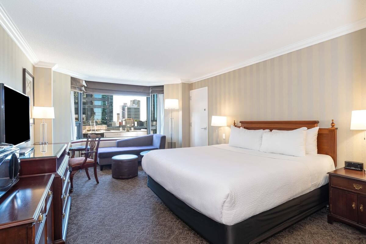 A hotel room with a large bed, a desk with a TV, a chair, a sofa, and a view of the city.