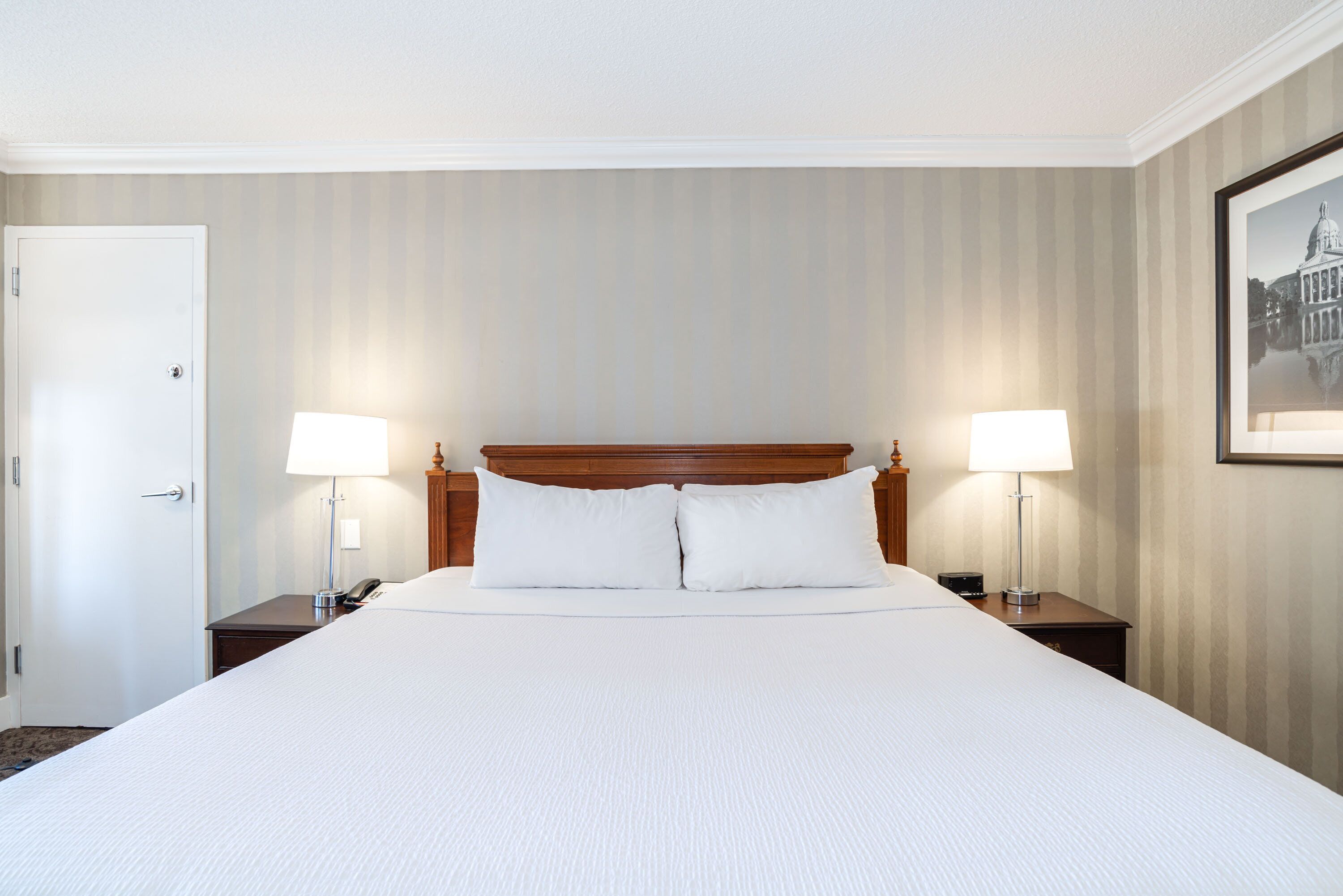 A large bed with white linens, two bedside tables with lamps, a wooden headboard, and a framed picture on the wall.