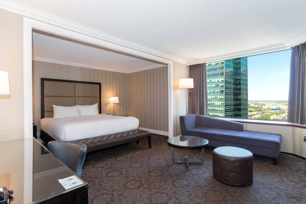 A hotel room with a large bed, a sofa, a small table, and a view of a cityscape through a large window.