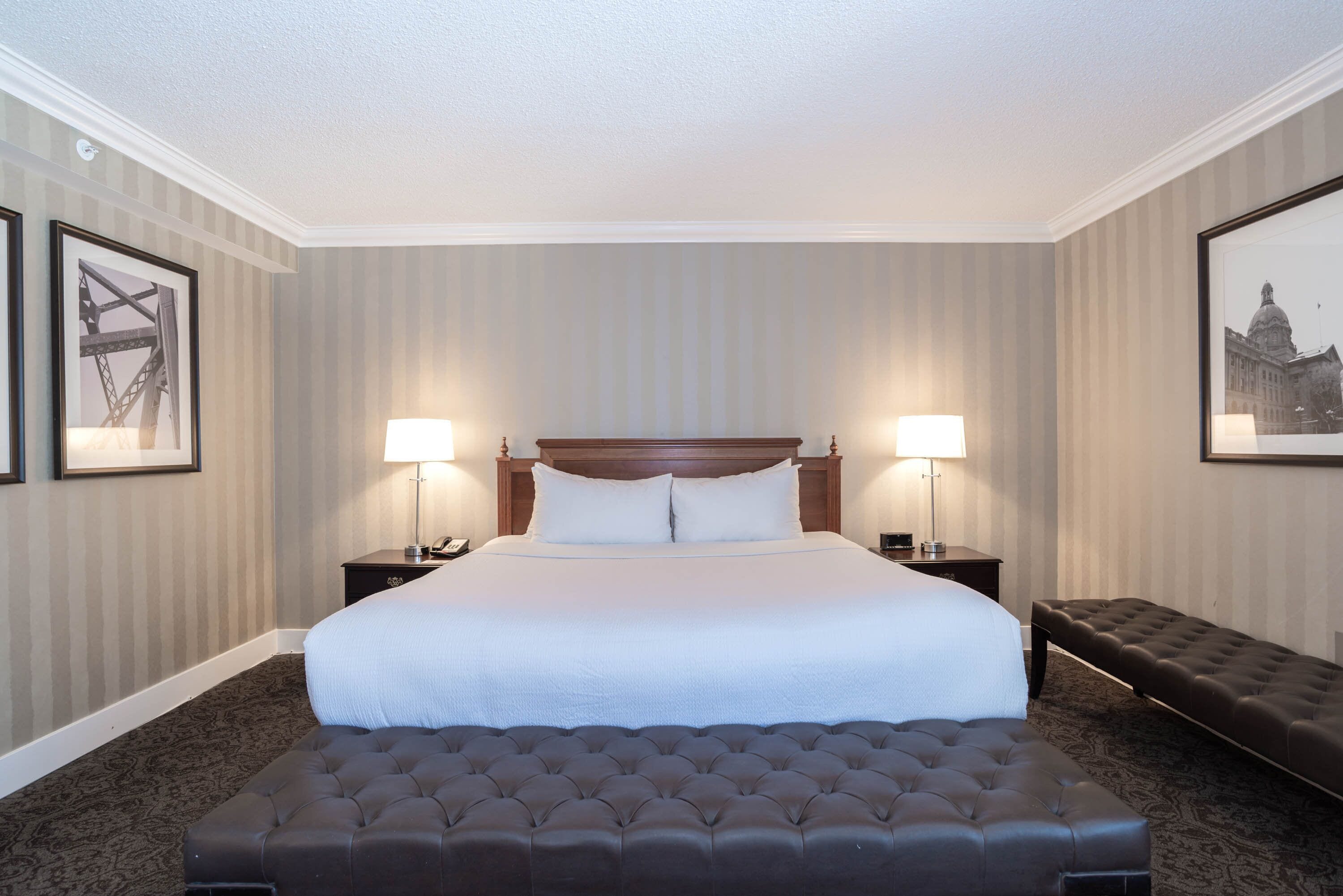 A hotel room with a large bed, two bedside tables with lamps, a dark upholstered bench, and framed pictures on the walls.
