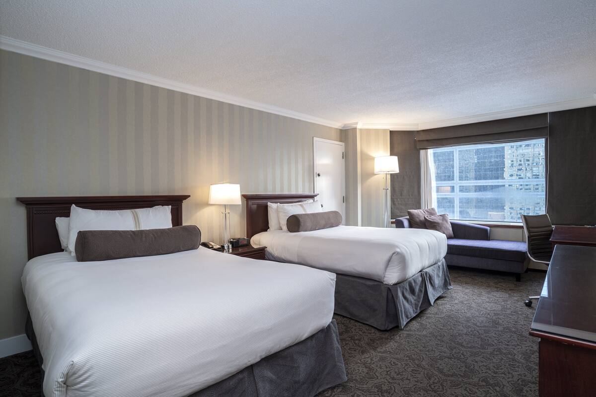 A hotel room with two beds, a desk, a chair, a window with a city view, and a door.
