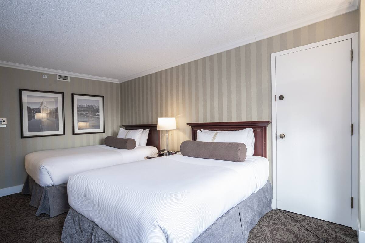 A hotel room with two beds, a nightstand, a lamp, and a door.