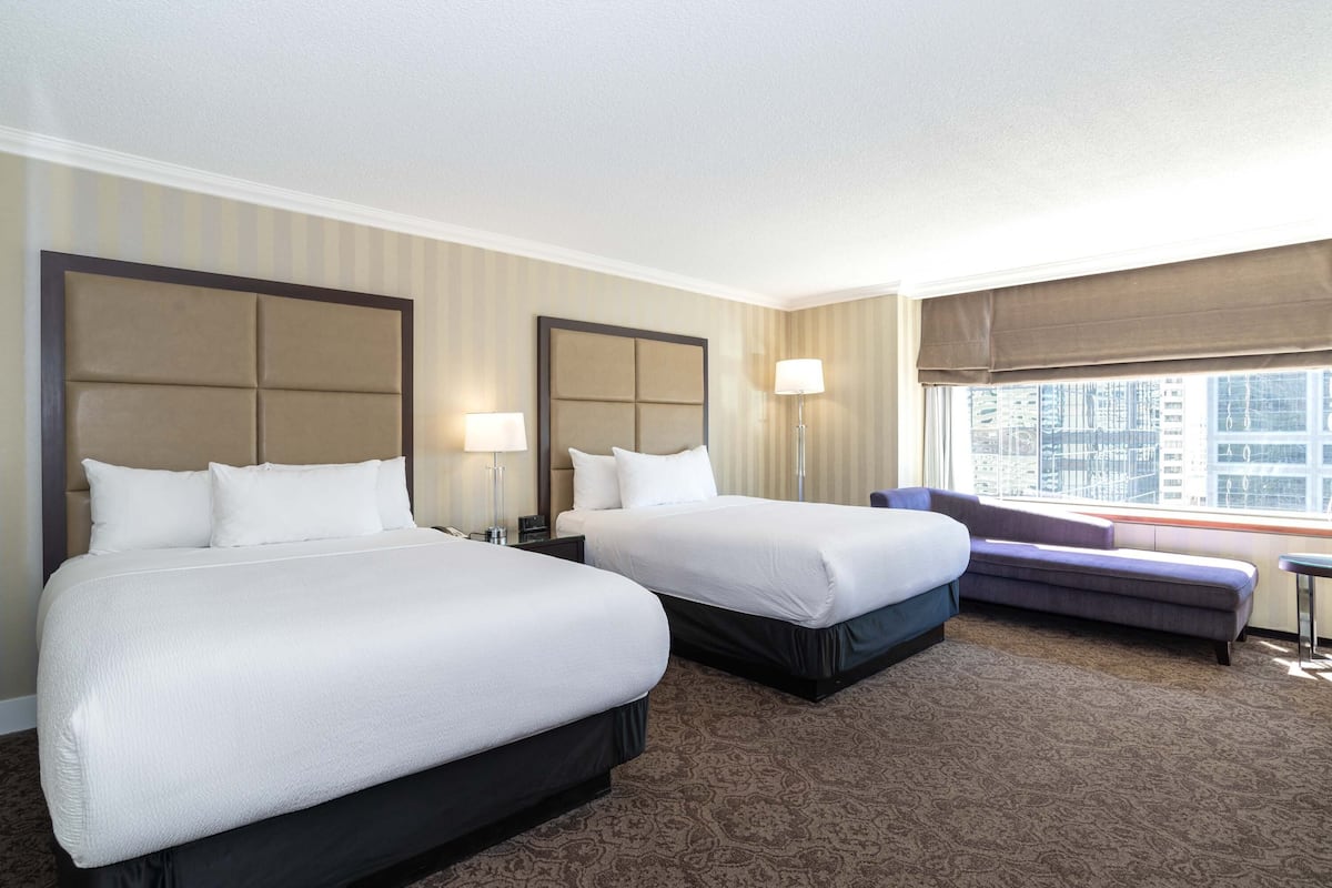 A hotel room with two beds, a sofa, a window with a view of buildings, and a patterned carpet.