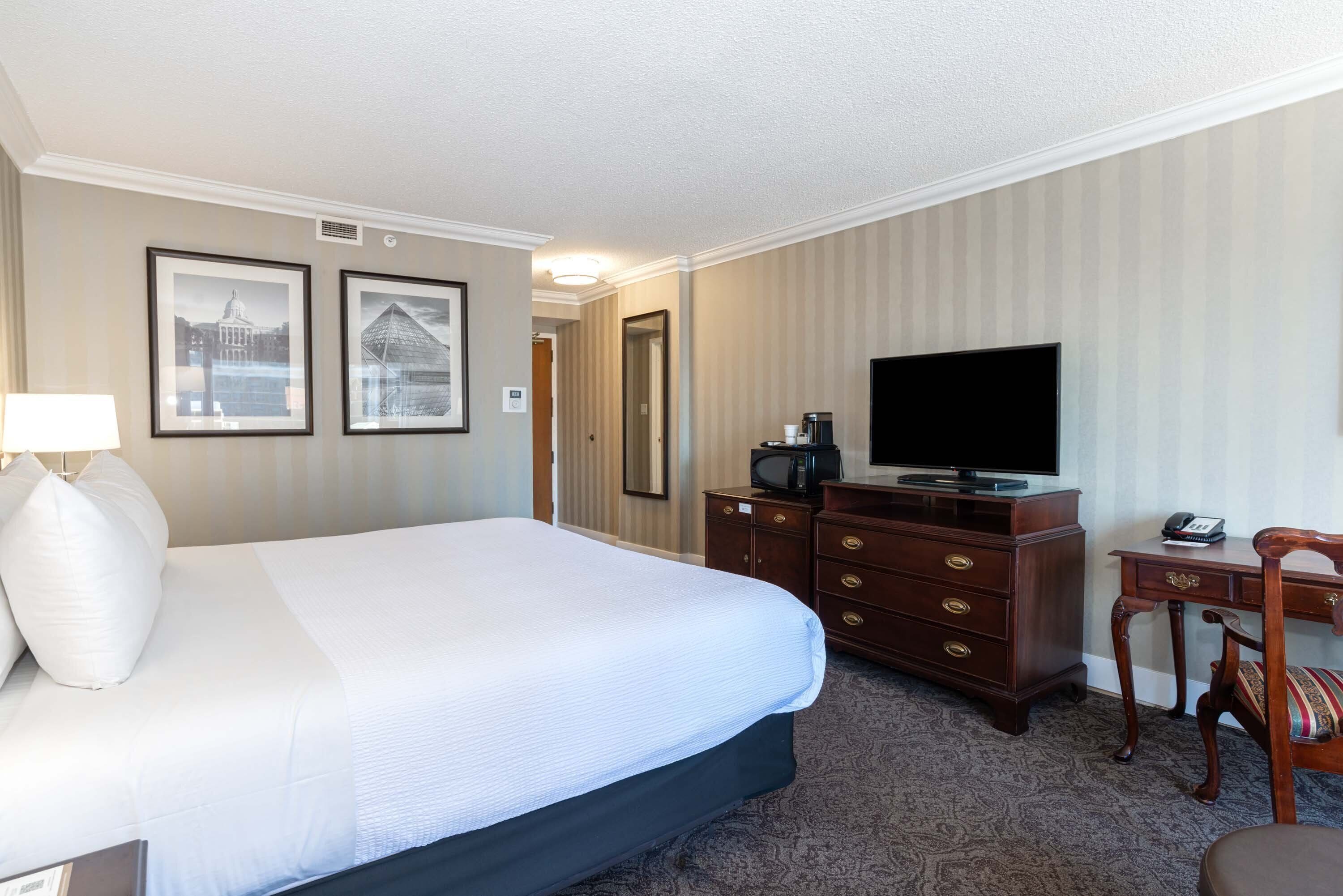 Habitación de hotel con una cama grande, un escritorio con televisor, un aparador y dos cuadros en la pared.