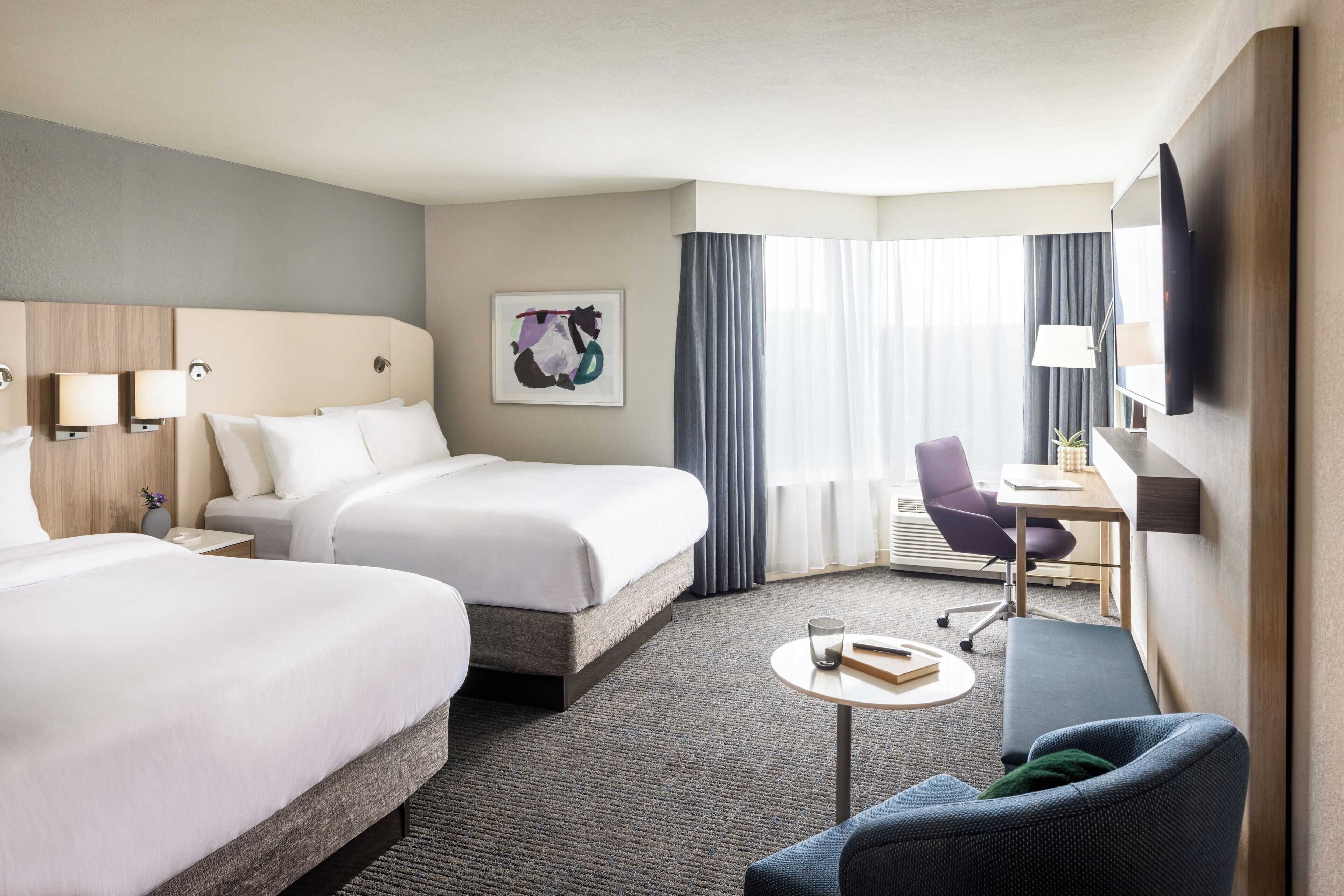 A hotel room with two beds, a desk, a chair, a TV, and a painting on the wall.