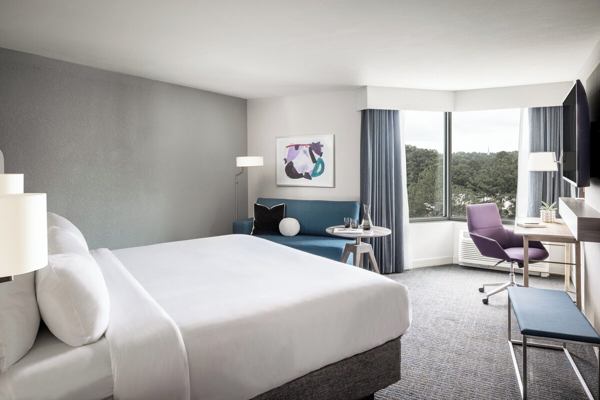 A hotel room with a large bed, a desk, a chair, a small table, and a large window offering a view of the outdoors.