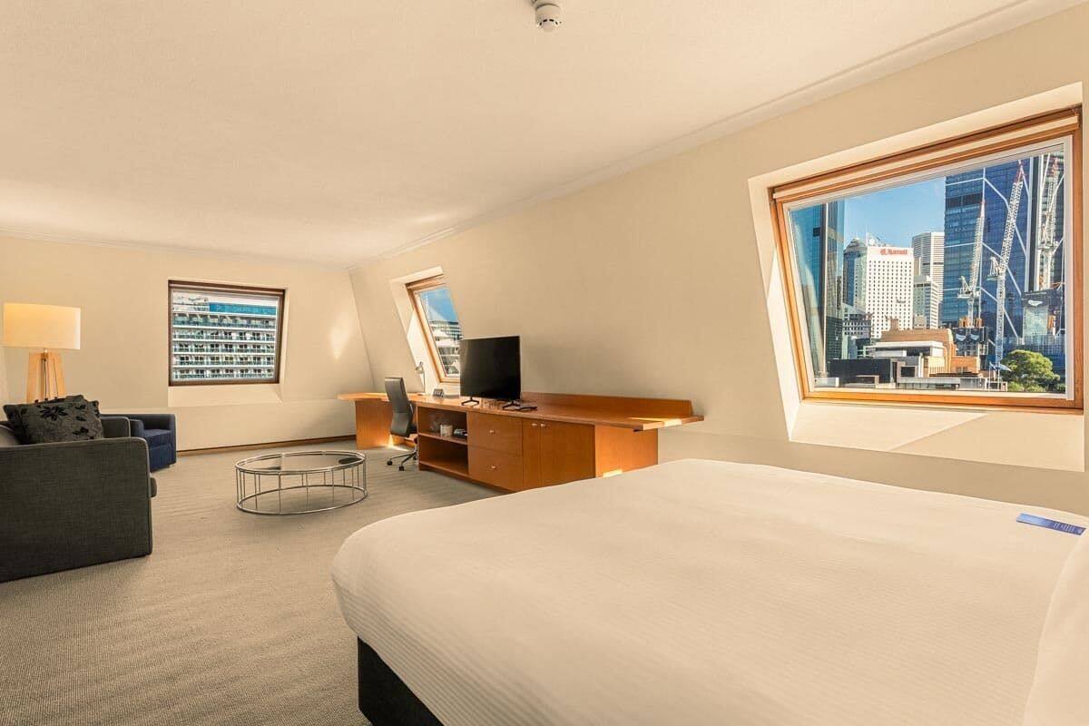 A hotel room with a large bed, a desk with a computer, a chair, a sofa, a lamp, and a view of a cityscape through a large window.
