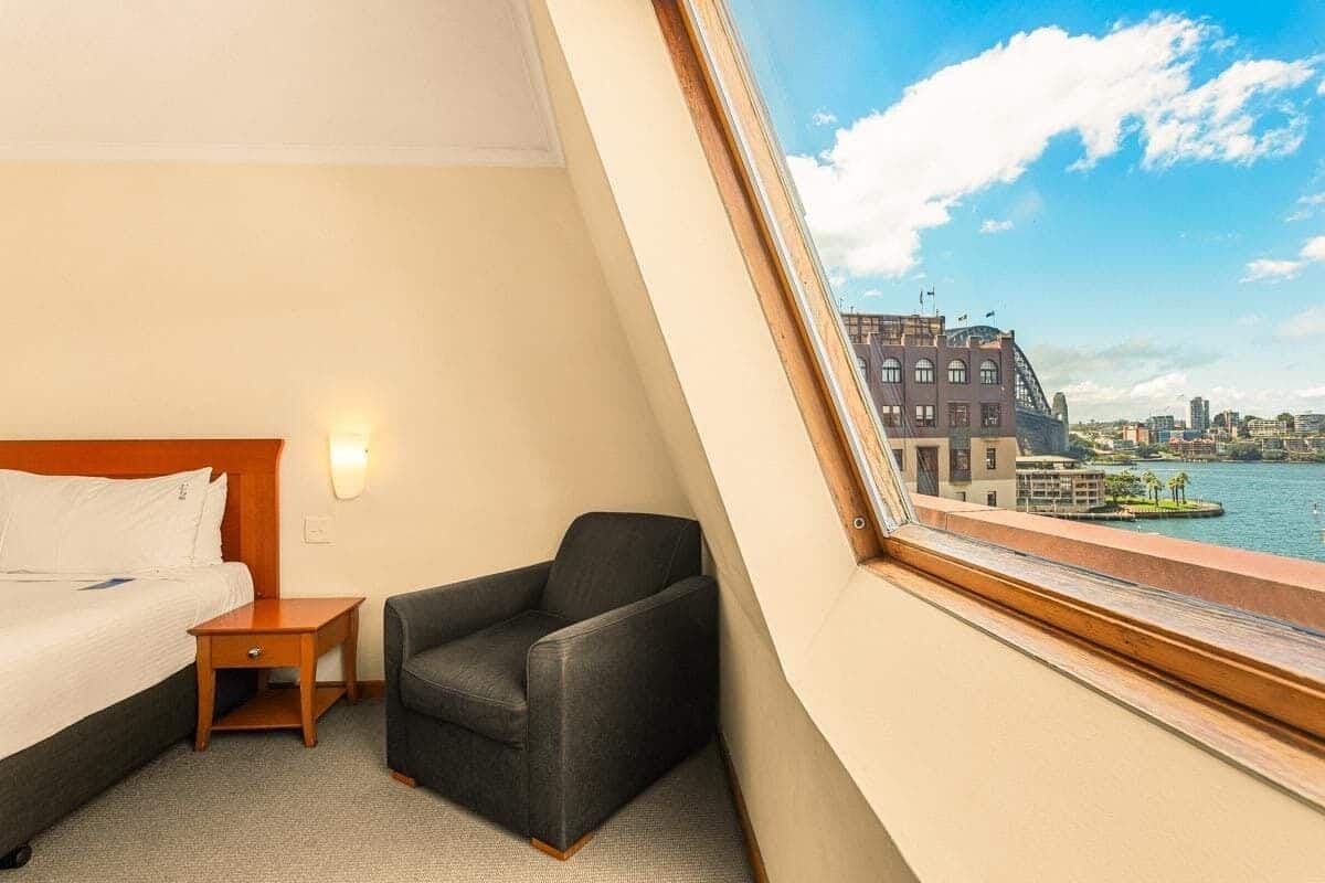 A hotel room with a bed, a chair, a nightstand, and a window offering a view of a cityscape and water.