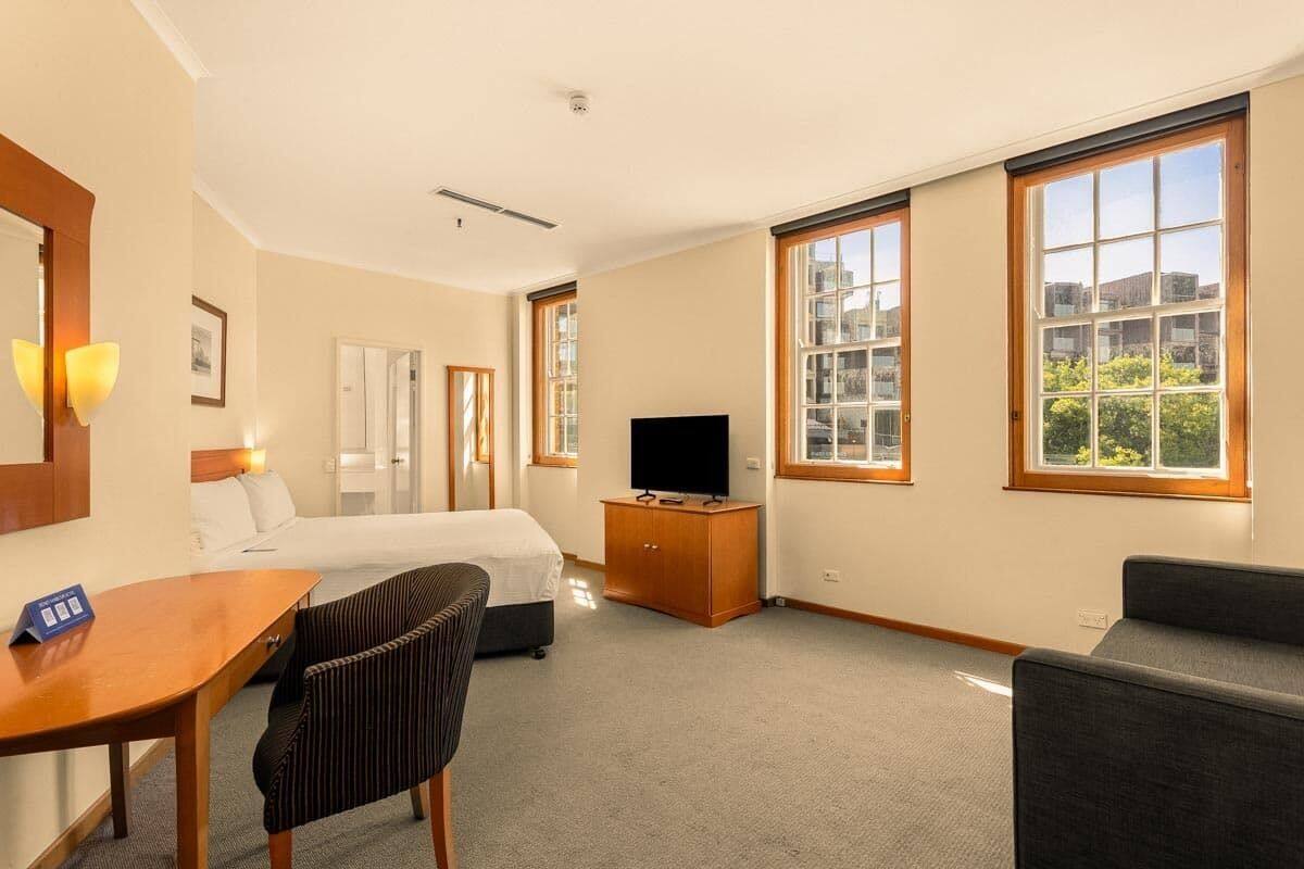 A hotel room with a bed, a desk, a television, and large windows.