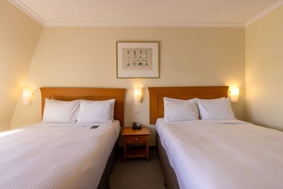 A hotel room with two beds, each with white linens and headboards, a small wooden nightstand with a phone, and a framed artwork on the wall.