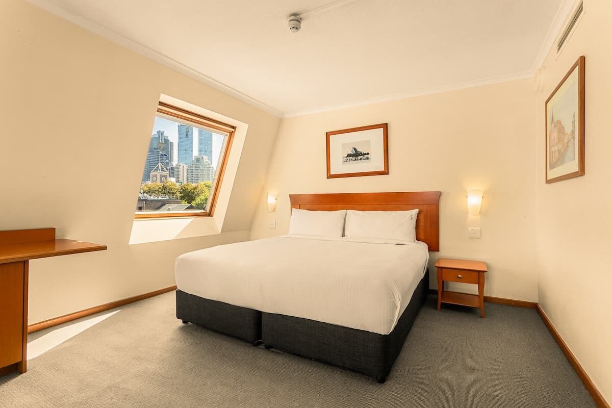 A hotel room with a bed, a desk, a nightstand, a window with a city view, and a framed picture on the wall.