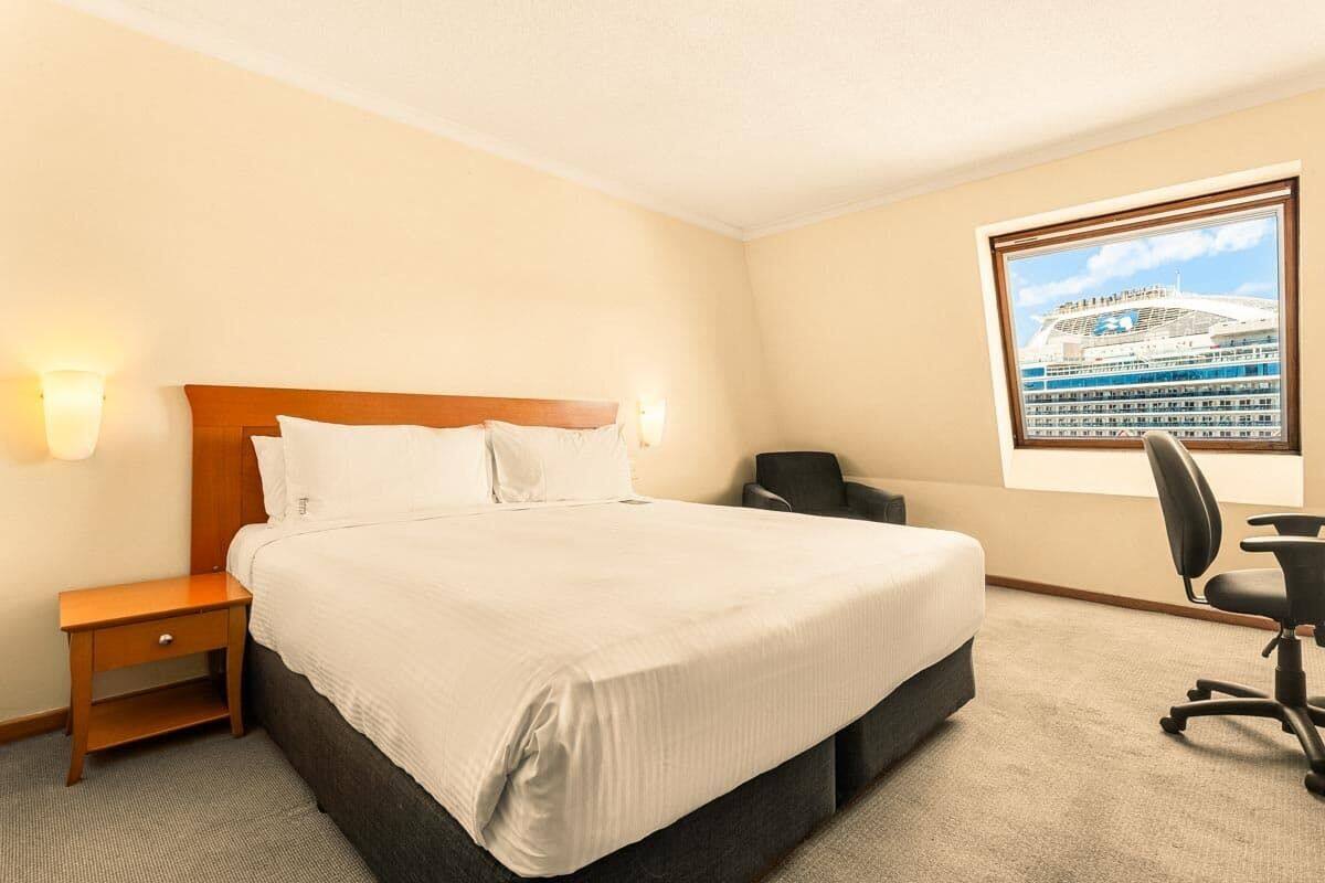 A hotel room with a large bed, a desk, and a window with a view of a cruise ship.