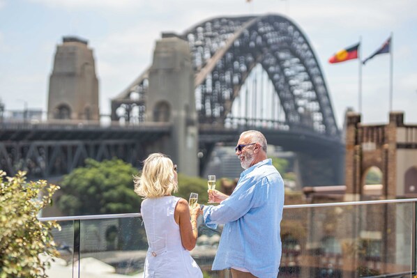 A couple standing on a balcony with a view of a large bridge and flags.