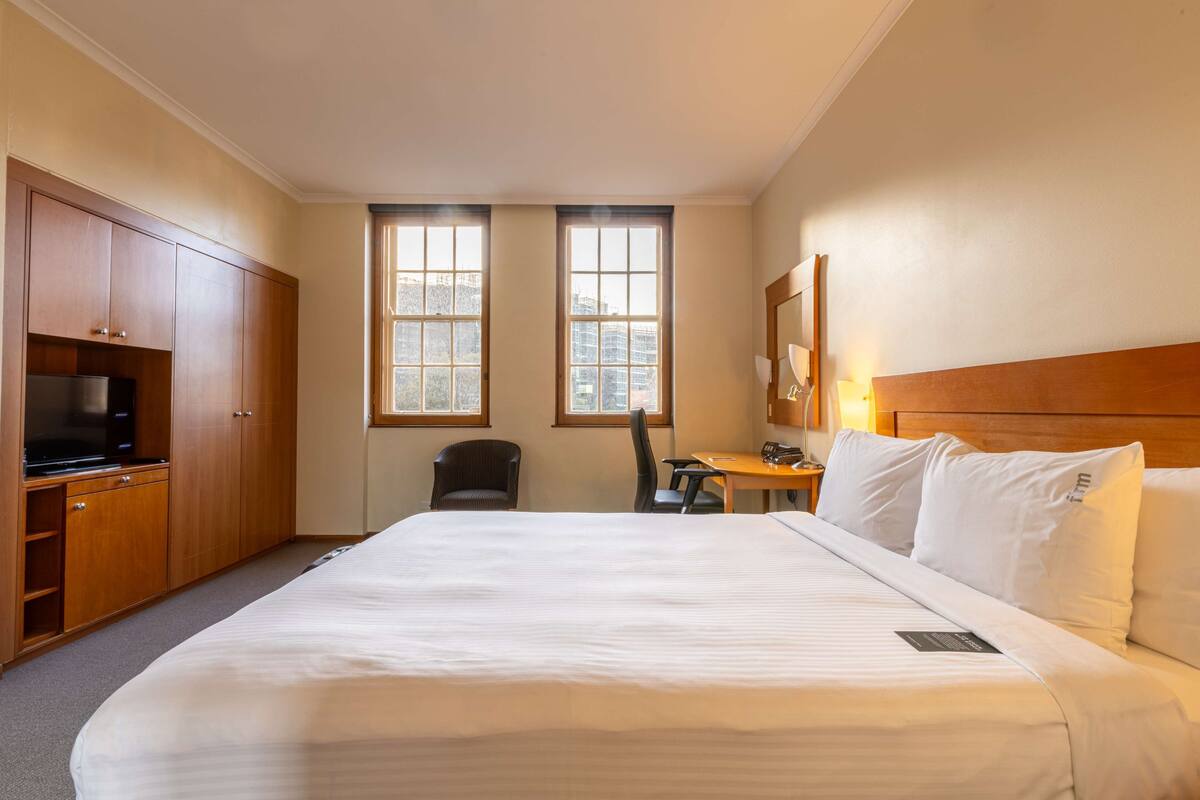 A hotel room with a large bed, wooden wardrobe, desk, chair, and two windows with city views.