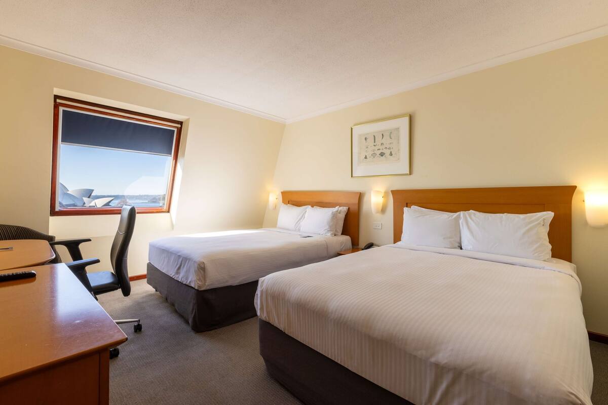 A hotel room with two single beds, a desk, a chair, and a window with a view of buildings.