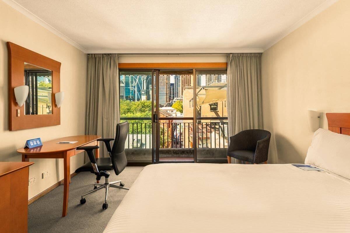 A hotel room with a large bed, a desk, a chair, and a balcony with a view of the city.