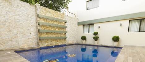 A swimming pool with a clear blue water surface, surrounded by a light-colored tiled deck and a stone wall.