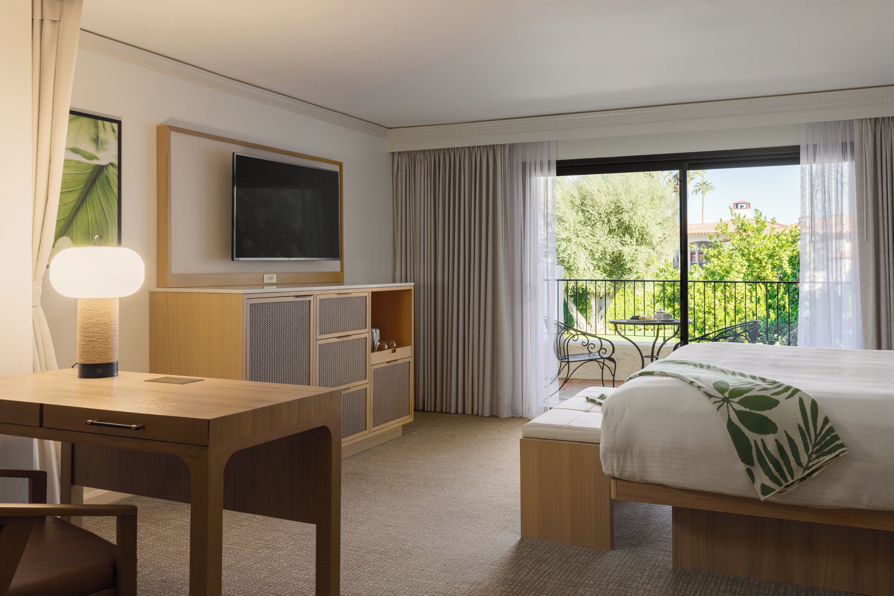A hotel room with a bed, a desk, a TV, and a balcony with a view of greenery and buildings.