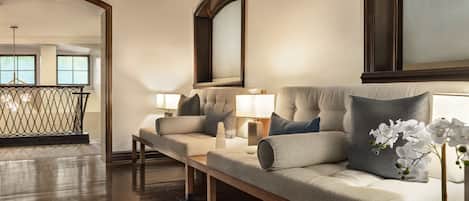 A spacious living room with a sofa set, a coffee table, and a mirror. There is a wooden floor and a hanging light fixture.