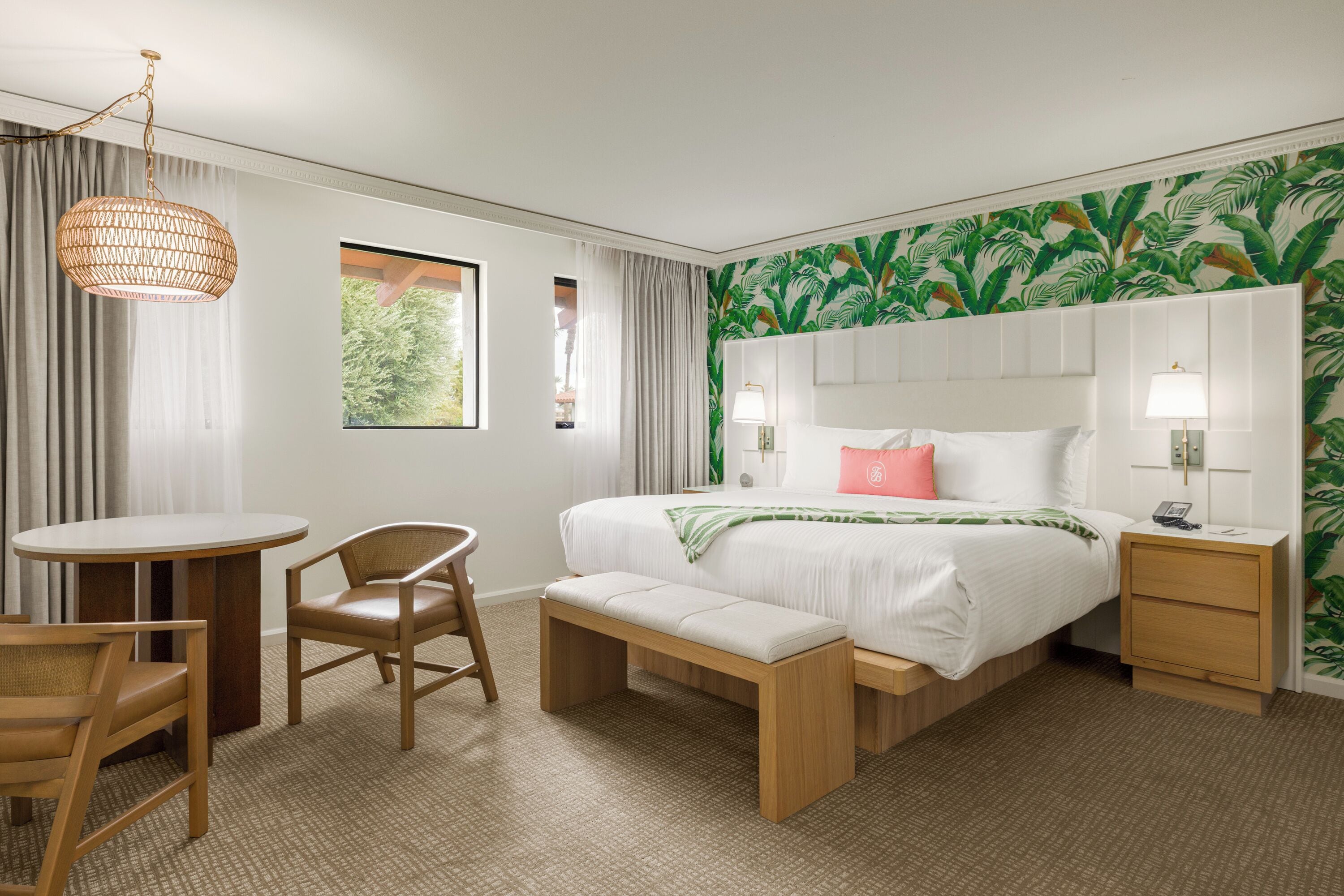 A hotel room with a bed, bedside table, lamp, chair, bench, and a view of greenery through the window.