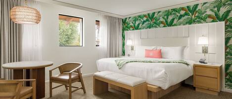 A hotel room with a bed, bedside table, lamp, chair, bench, and a view of greenery through the window.