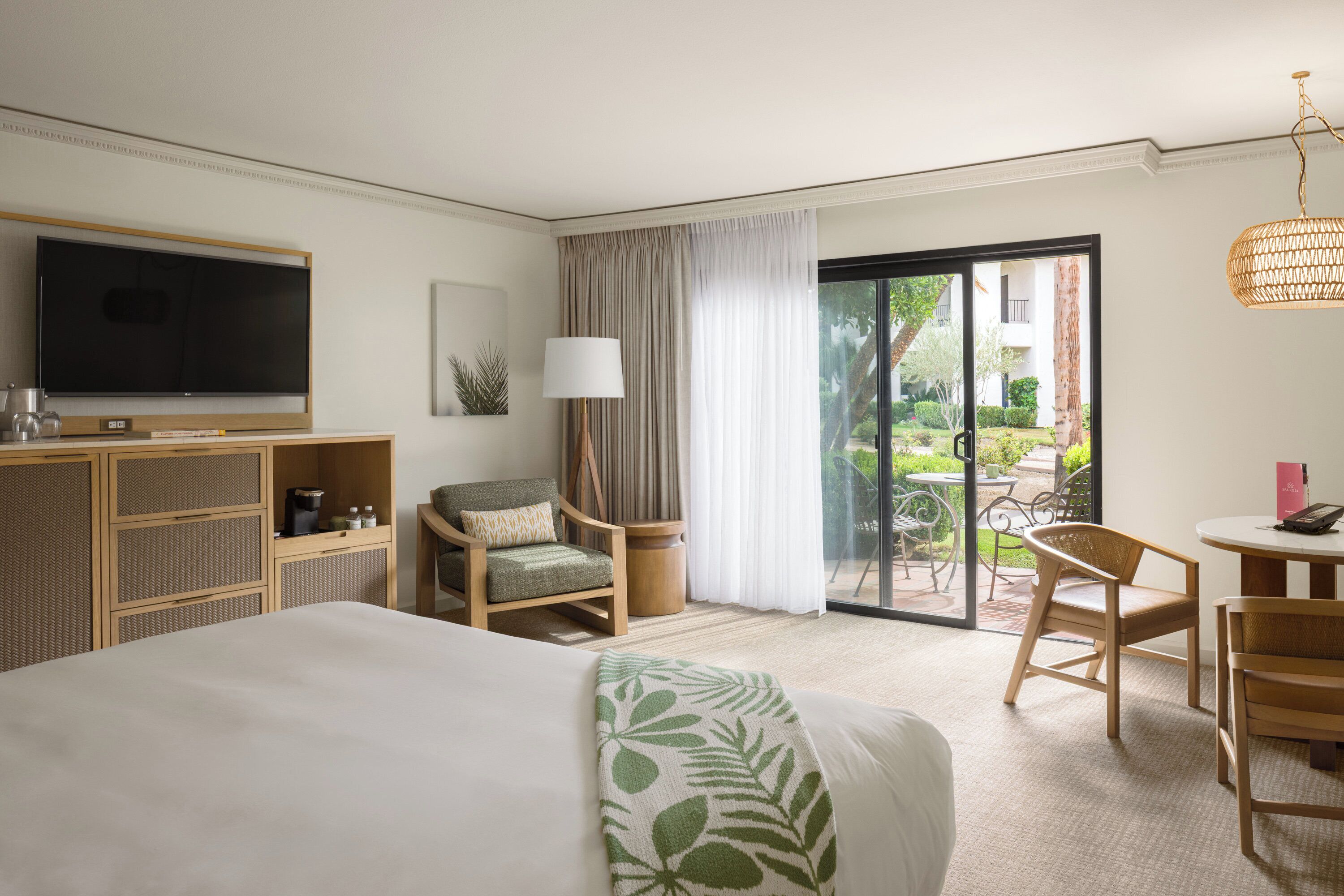 A hotel room with a bed, a television, a desk with a chair, a dining table with chairs, and a balcony with a view of the garden.