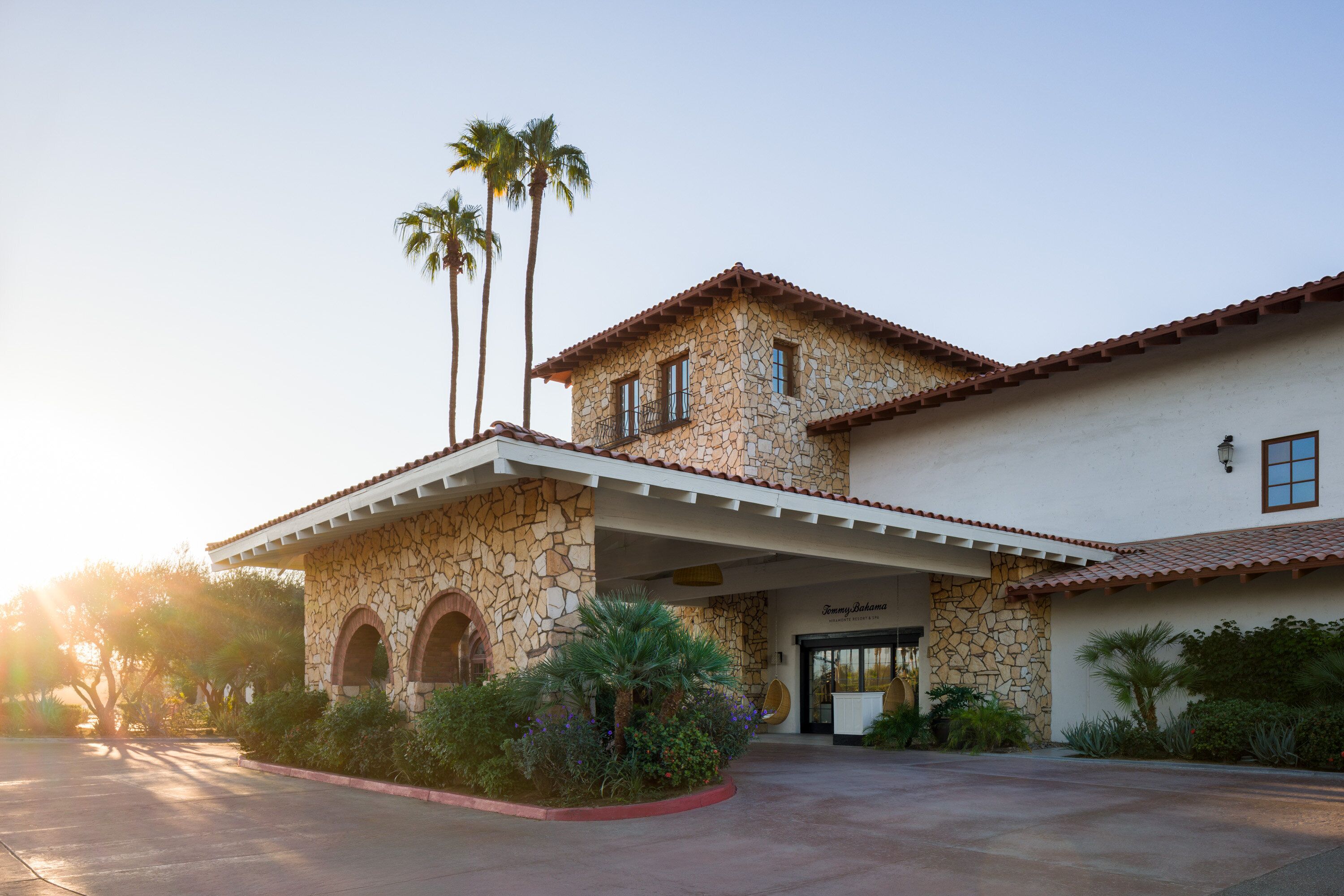 A two-story stone building with a covered patio, palm trees, and a clear sky.