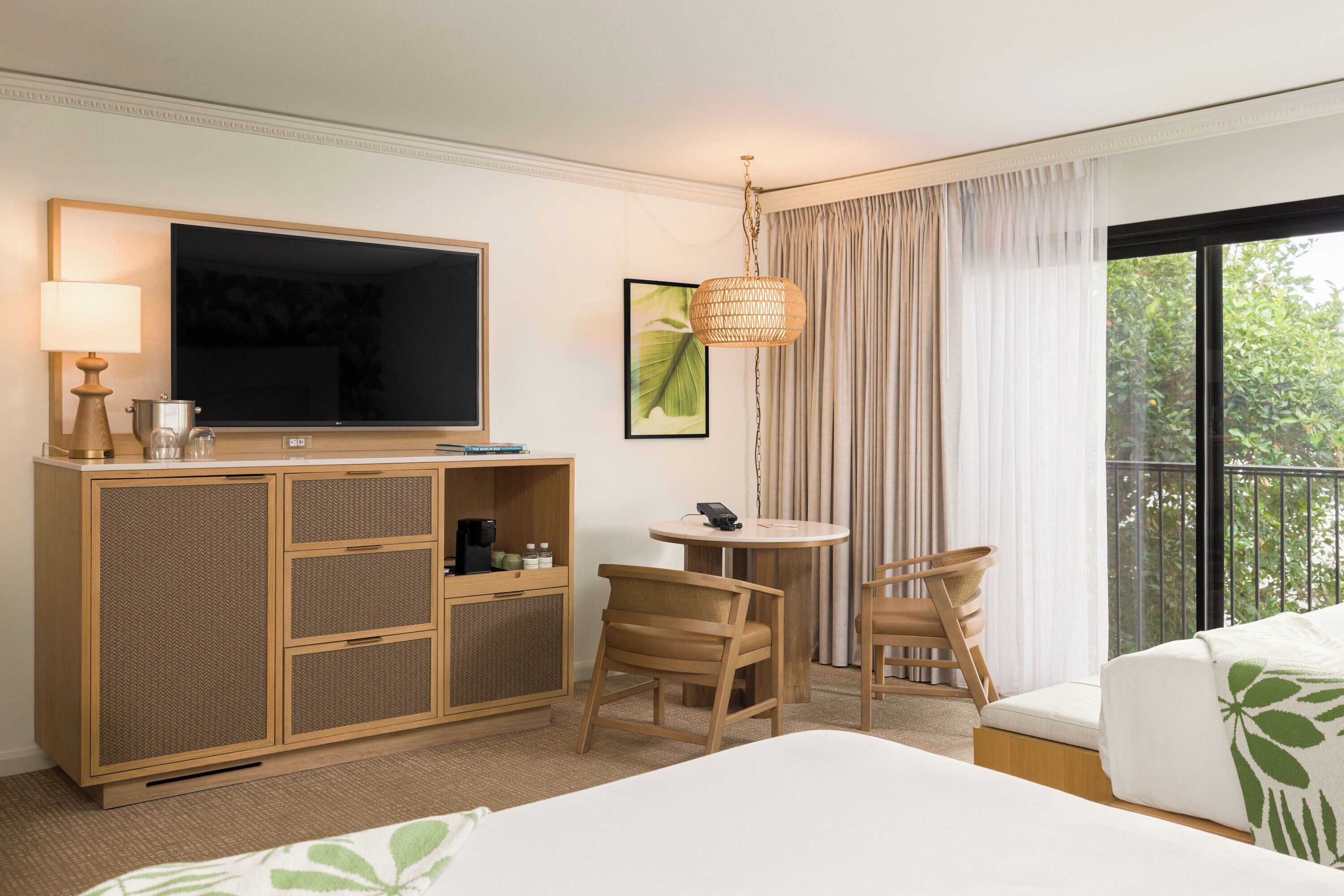 A hotel room with a bed, a TV, a desk with a lamp, a chair, a small table, and a balcony with curtains.
