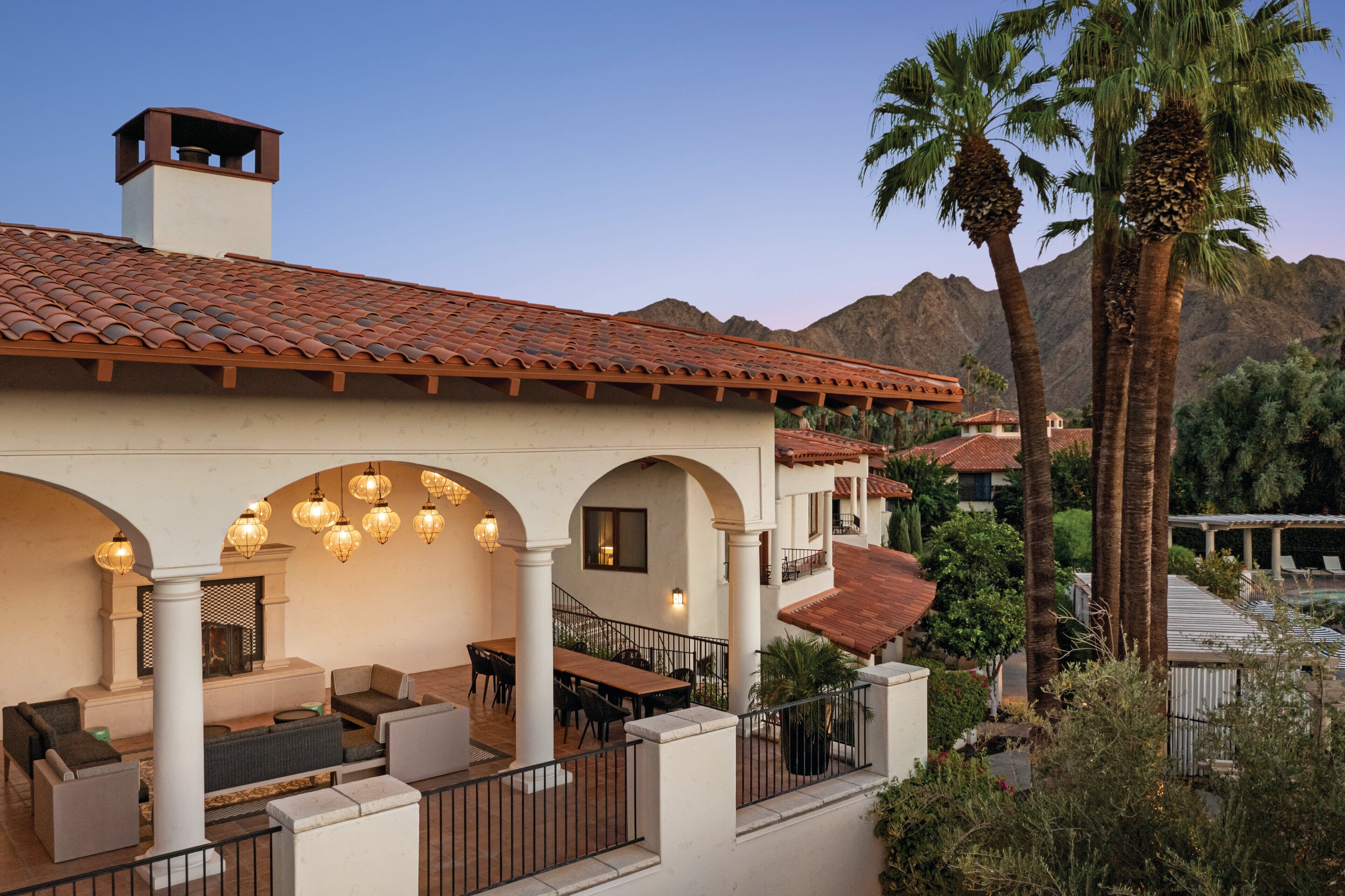 A Mediterranean-style building with a tiled roof, outdoor seating area with tables and chairs, and a view of palm trees and mountains in the background.
