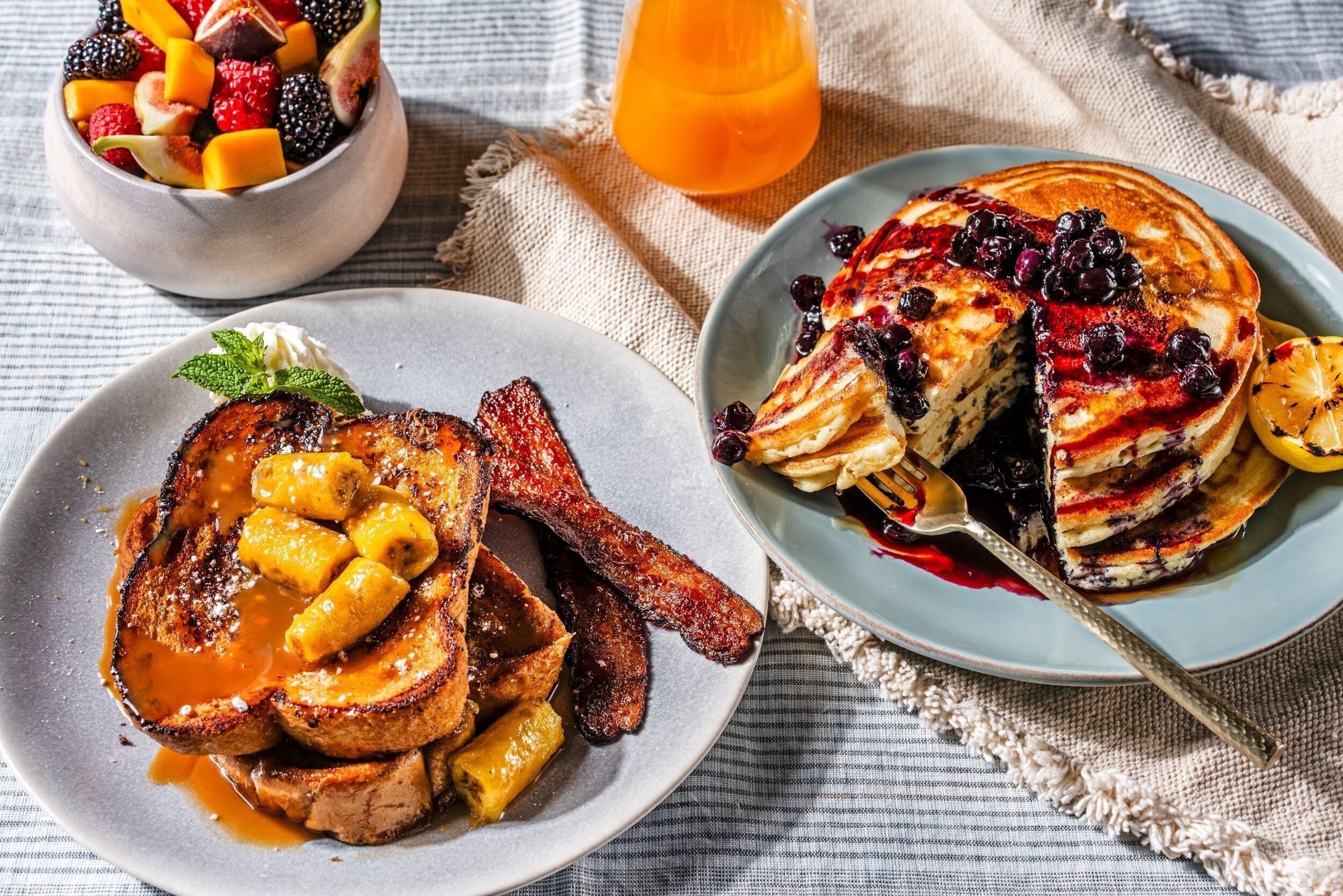 A breakfast setting with toast, bacon, and pancakes on plates, accompanied by a fruit bowl and a glass of orange juice.