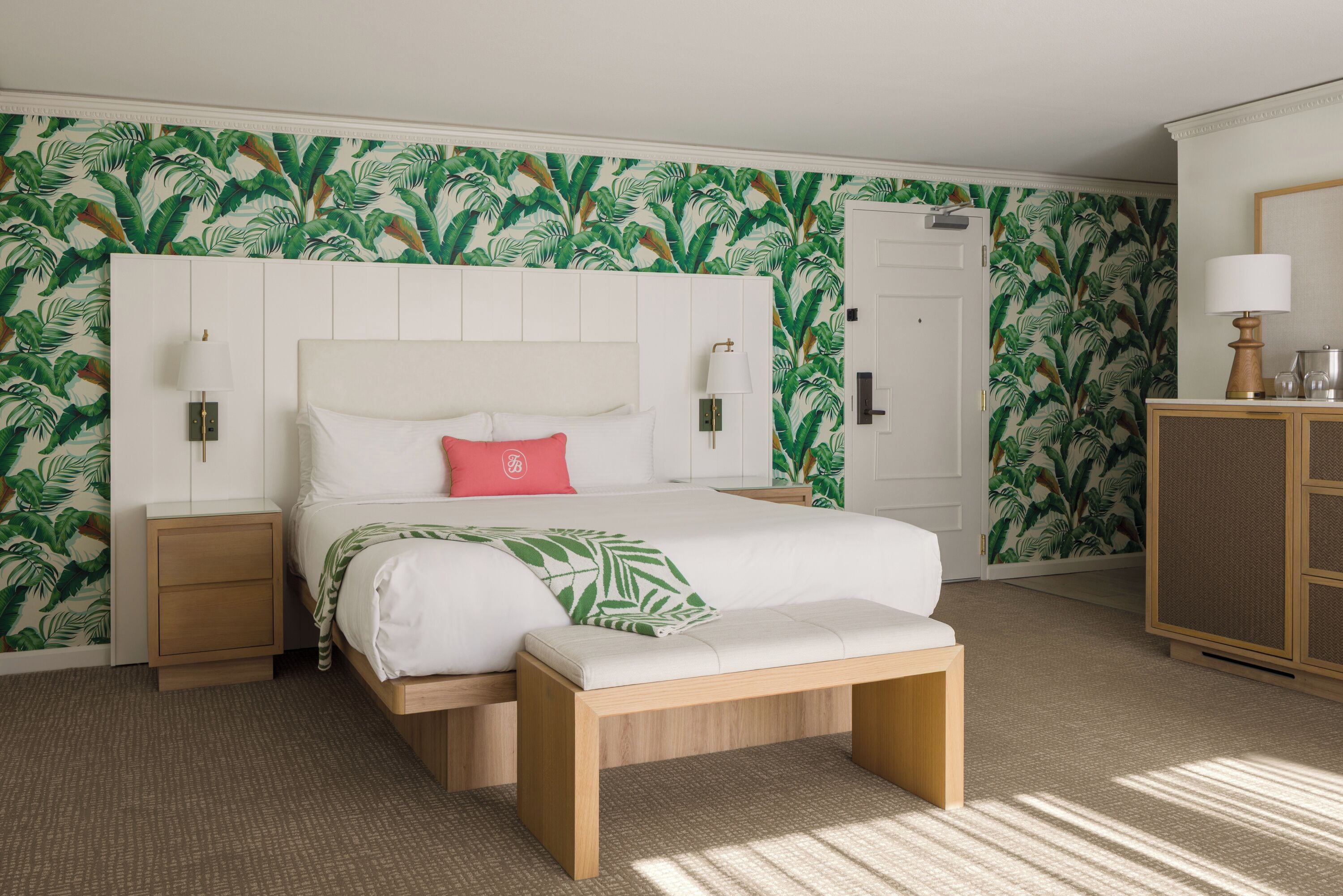 Una habitación de hotel con cama, banco, mesita de noche, cómoda y puerta.