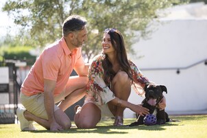 Una pareja sentada sobre la hierba con un perro, una tienda de campaña blanca y un árbol al fondo.