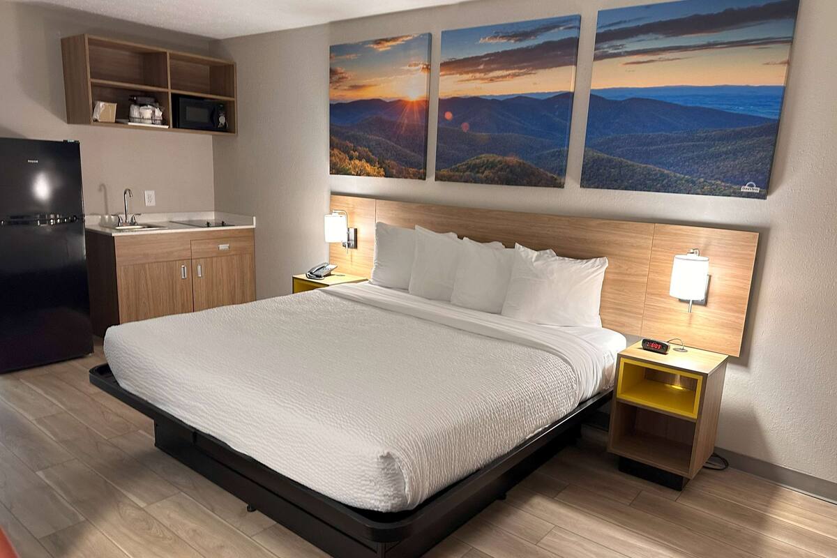 Une chambre d’hôtel comprenant un lit, une table de chevet, un bureau avec une lampe, un four à micro-ondes et une étagère avec divers objets.