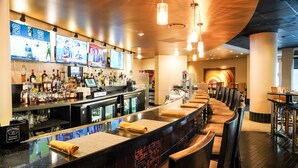 A bar area with a long counter, multiple screens displaying sports, a variety of liquor bottles, and a seating area with cushioned chairs. - Holiday Inn Washington Capitol - Natl Mall by IHG (Washington)