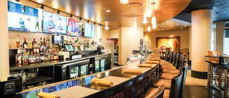 Un espace bar doté d’un long comptoir, de plusieurs écrans diffusant des événements sportifs, d’une grande variété de bouteilles d’alcool et d’un coin salon avec des fauteuils confortables.