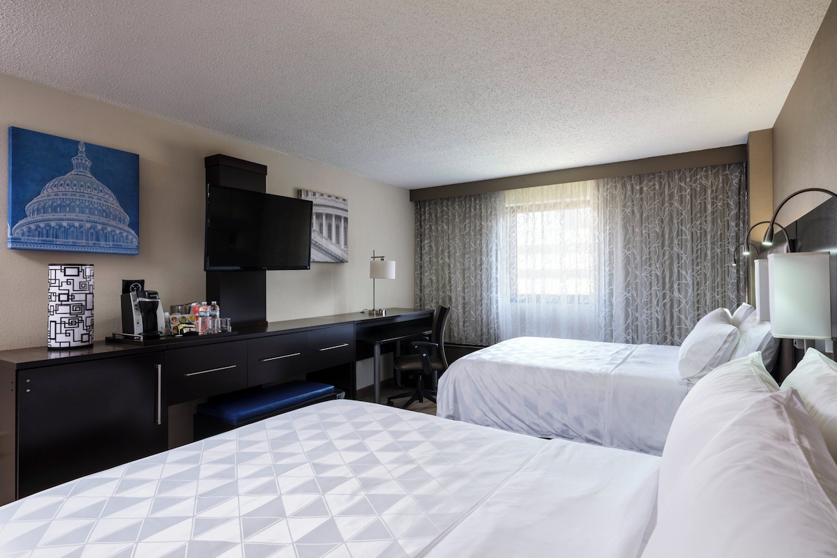 A hotel room with two beds, a desk, a television, and a painting of the Capitol.