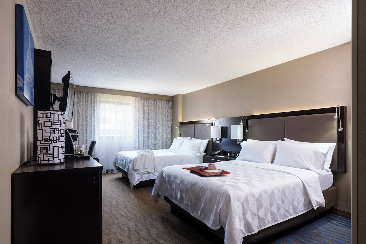 A hotel room with two beds, a desk, a TV, and a window with curtains.