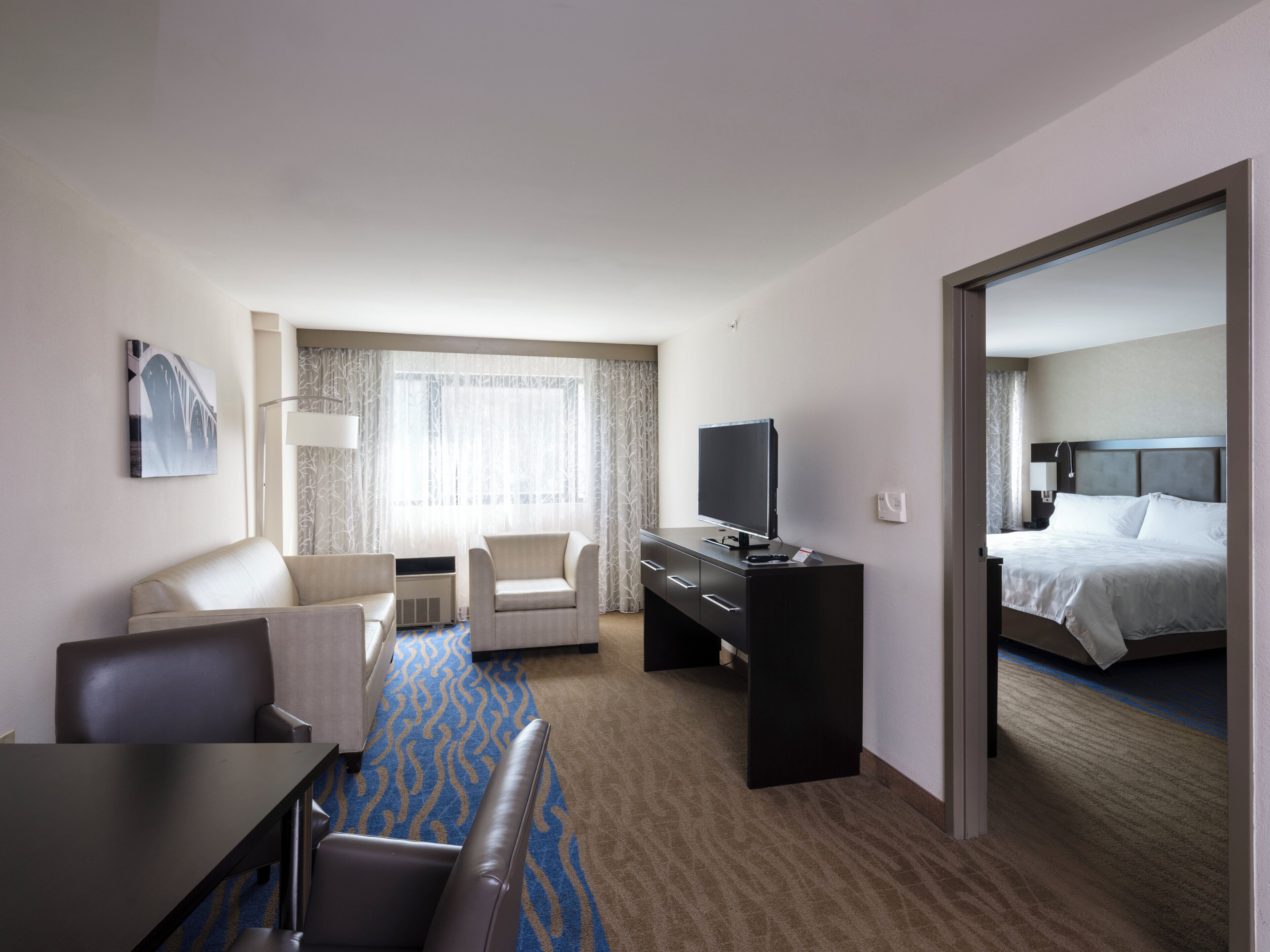 A hotel room with a bed, a desk with a TV, a sofa, a chair, a window with curtains, and a carpeted floor.