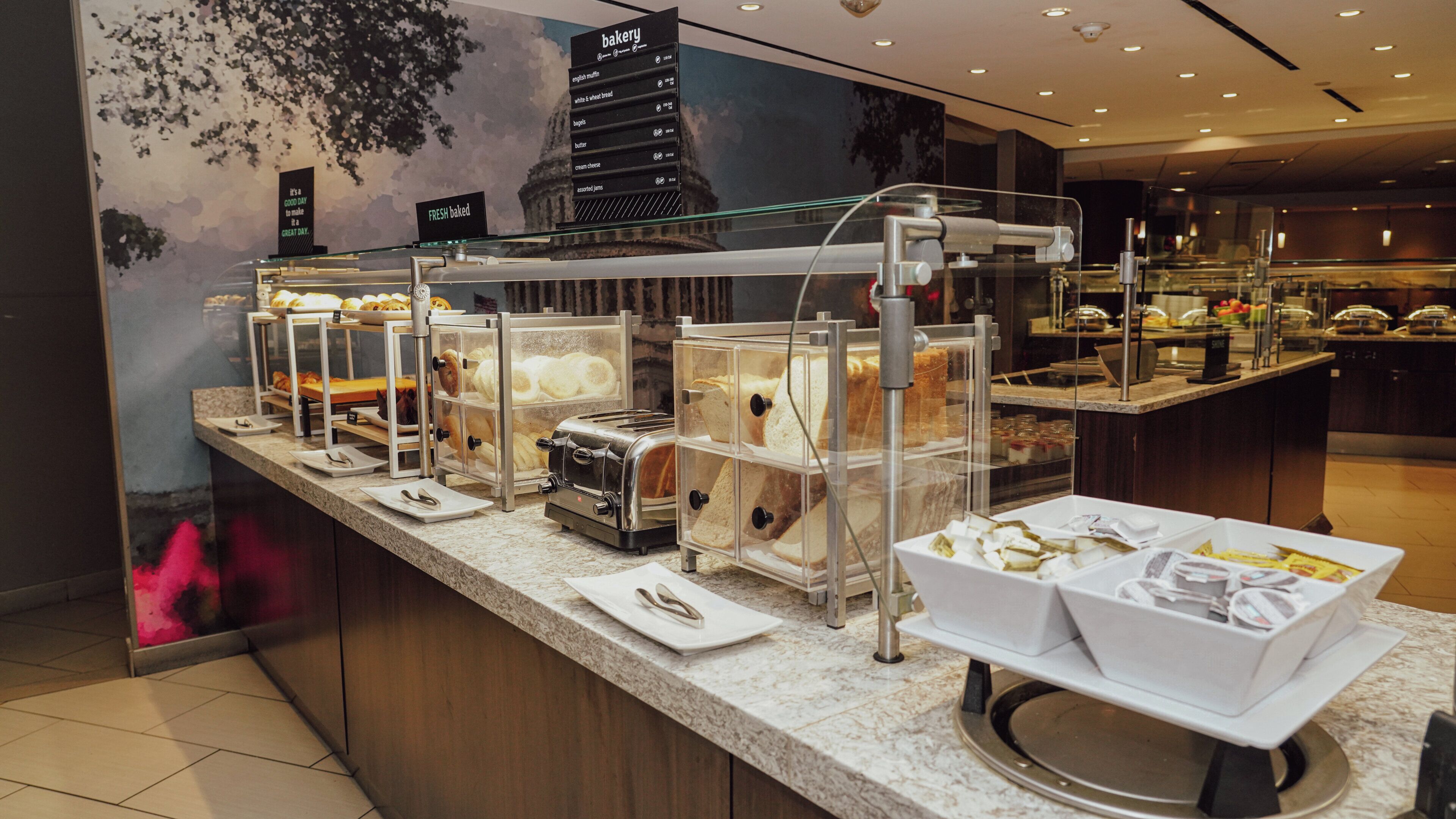 Mostrador de panadería con diversas opciones de productos horneados exhibidos en recipientes transparentes, un letrero que indica productos recién horneados y un panel con la carta de la panadería.