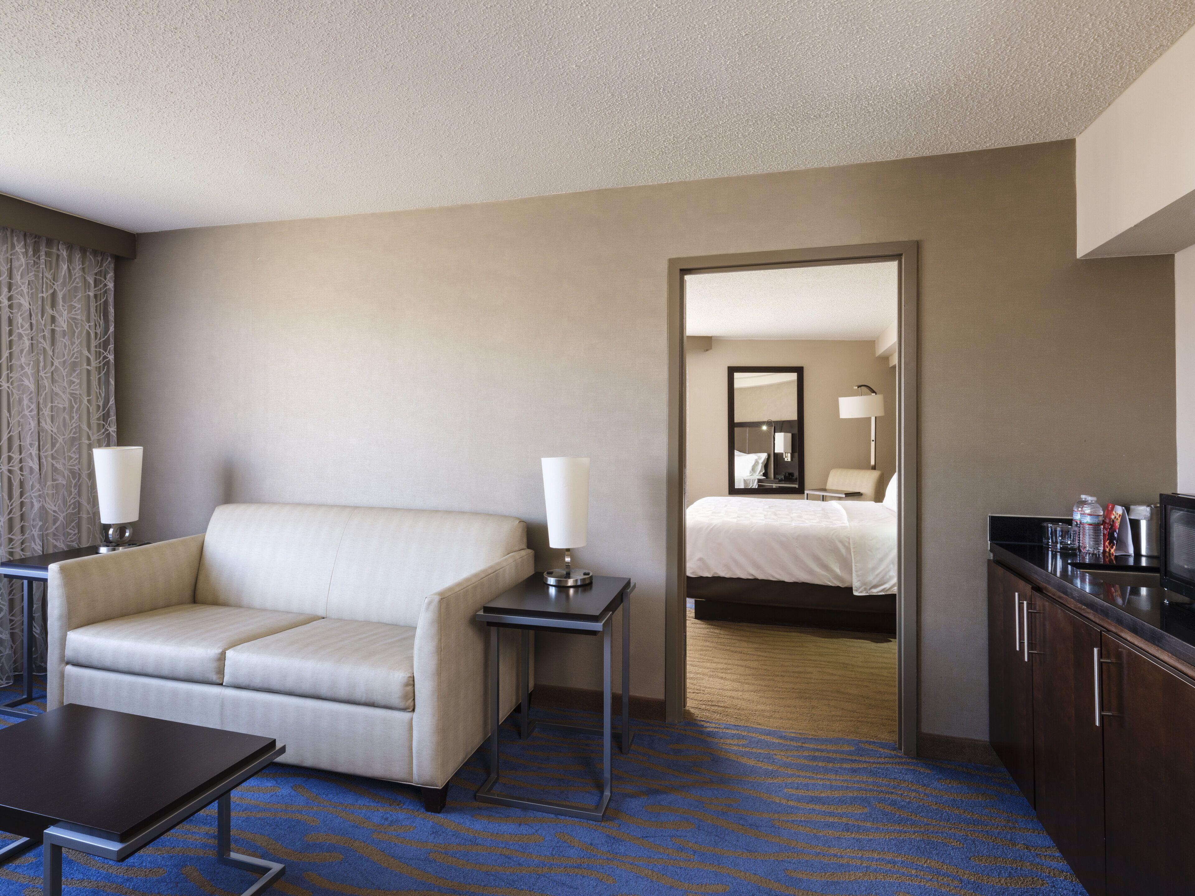 A hotel room with a sofa, a small table, a lamp, a bed, a mirror, and a minibar.