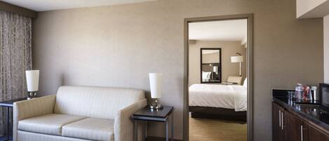 A hotel room with a sofa, a small table, a lamp, a bed, a mirror, and a minibar.