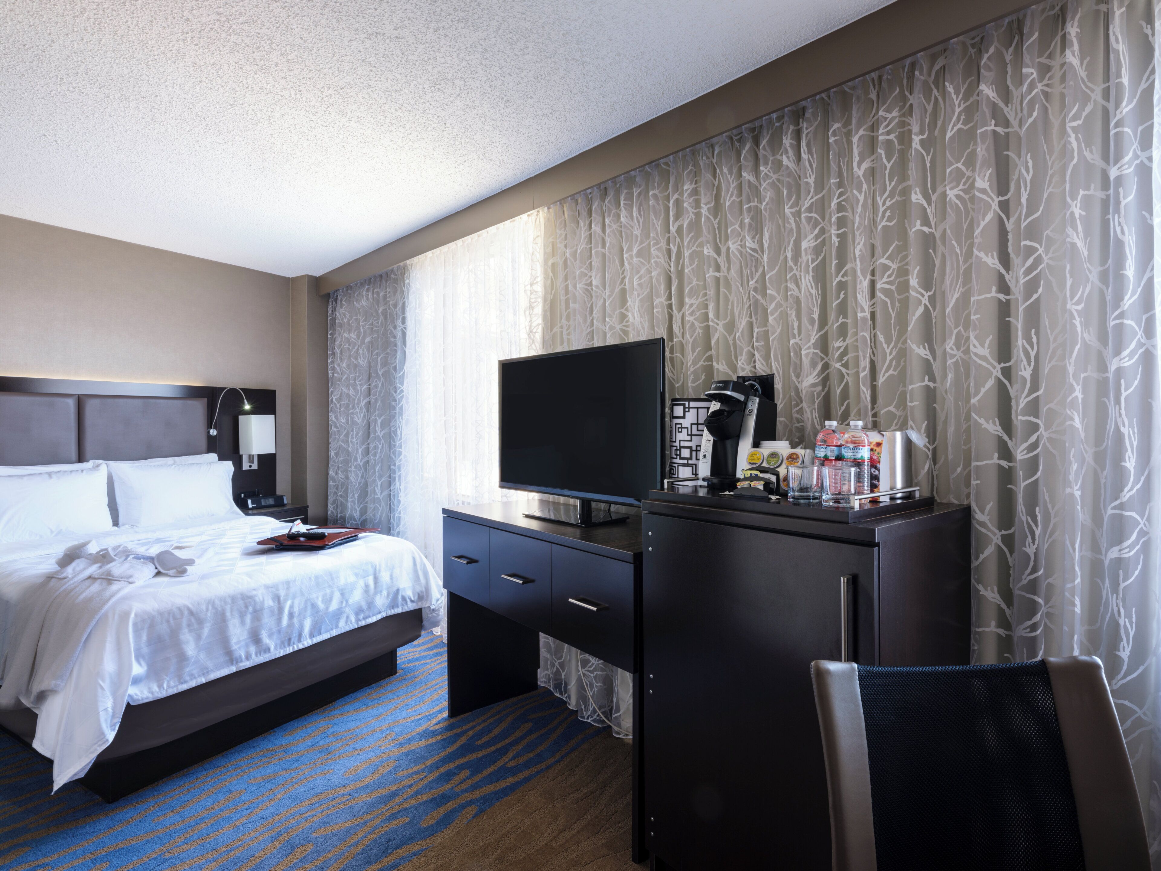 A hotel room with a bed, a desk with a TV, a coffee maker, and a chair.