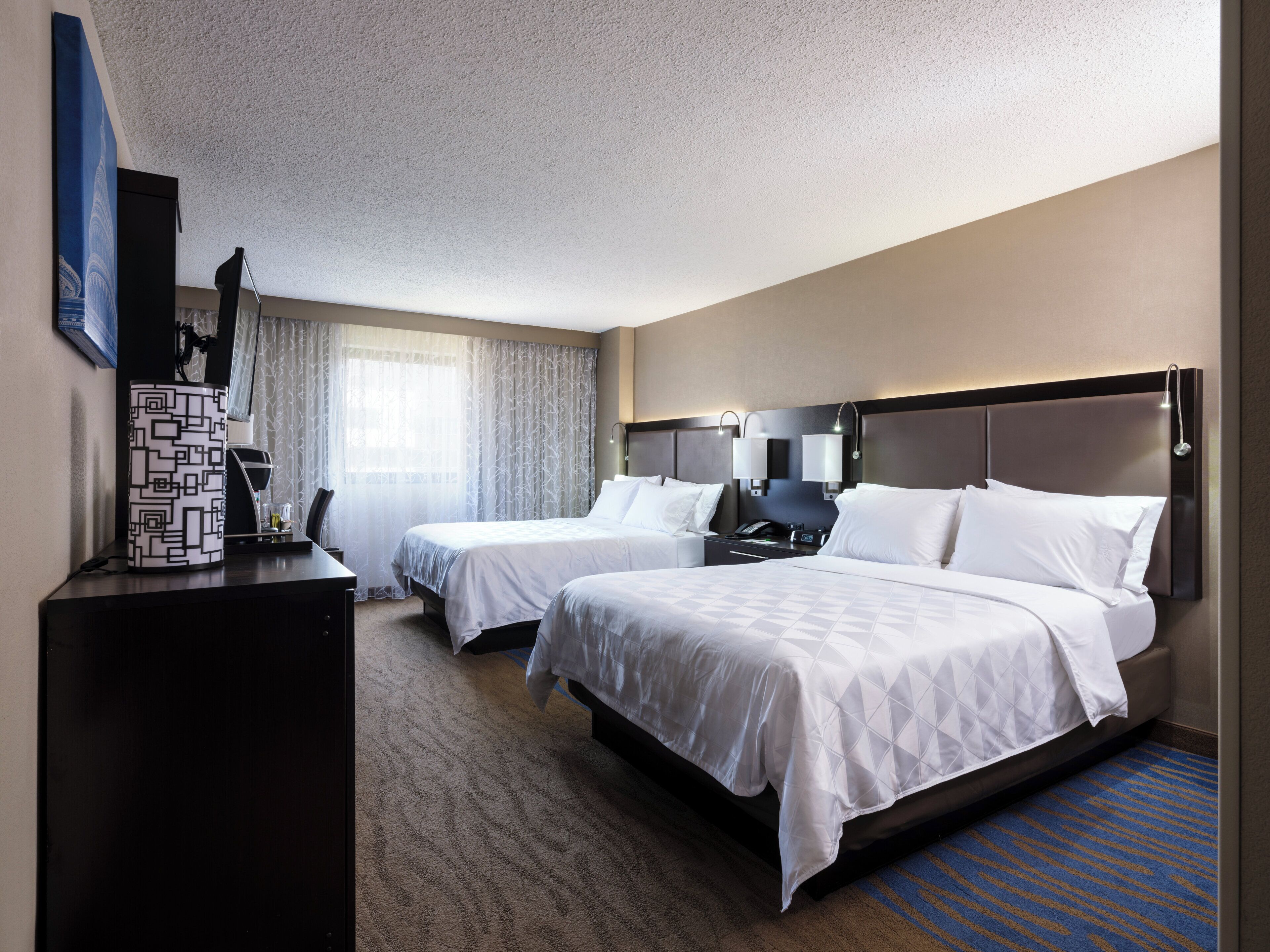 Quarto de hotel com duas camas, uma escrivaninha, uma cadeira, uma televisão e uma janela com cortinas.